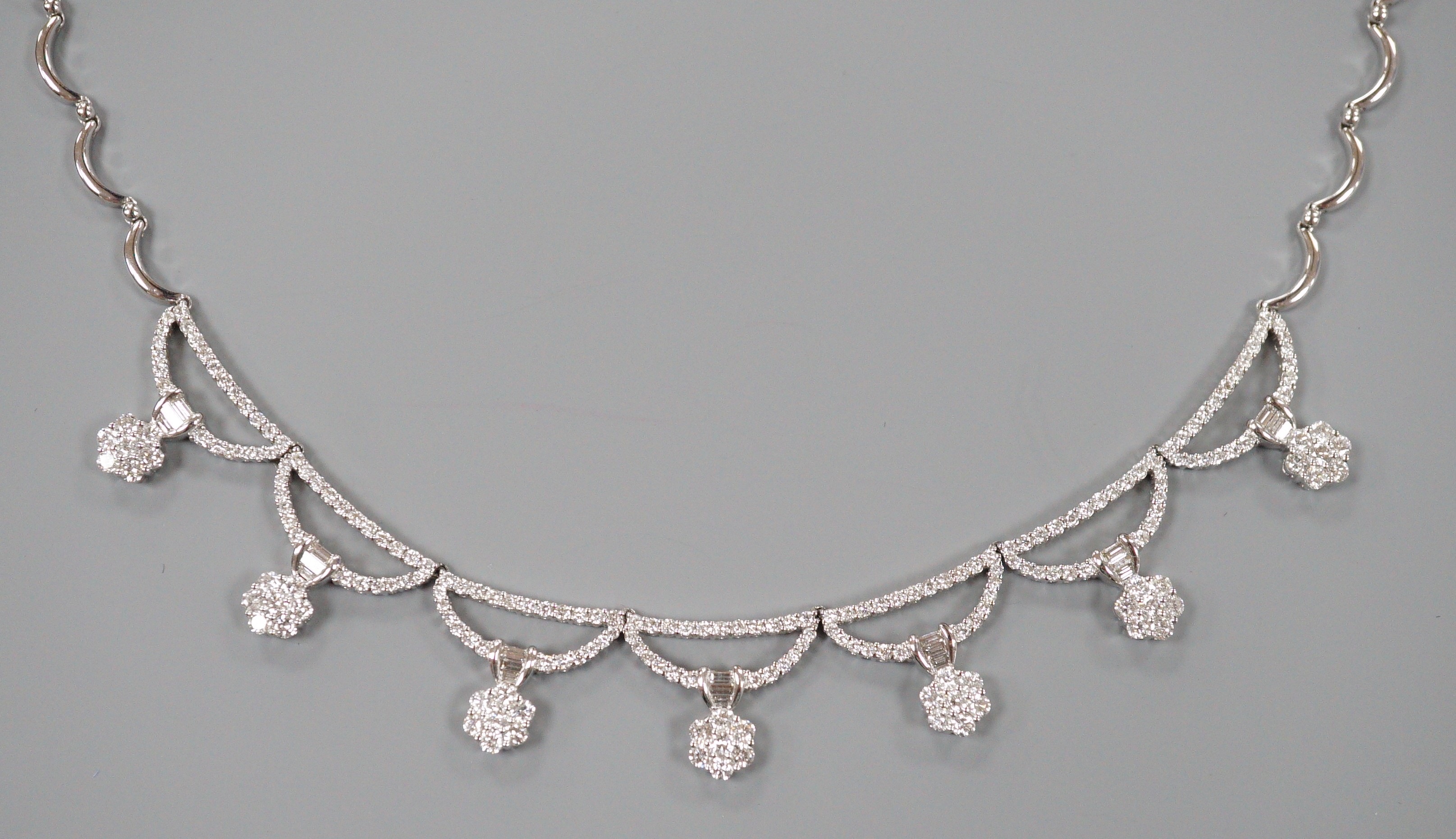 A modern 750 white metal and septuple baguette and round cut diamond cluster set curved link fringe necklace, 43cm, gross weight 26.6 grams